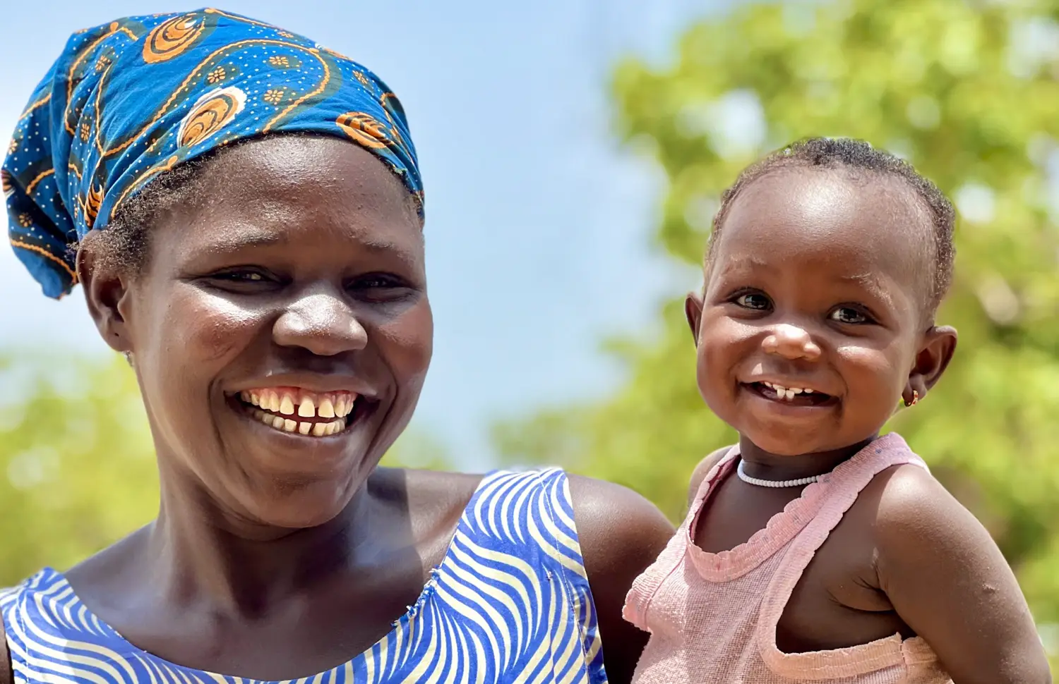A smiling mother in a blue headscarf holds her young daughter outdoors