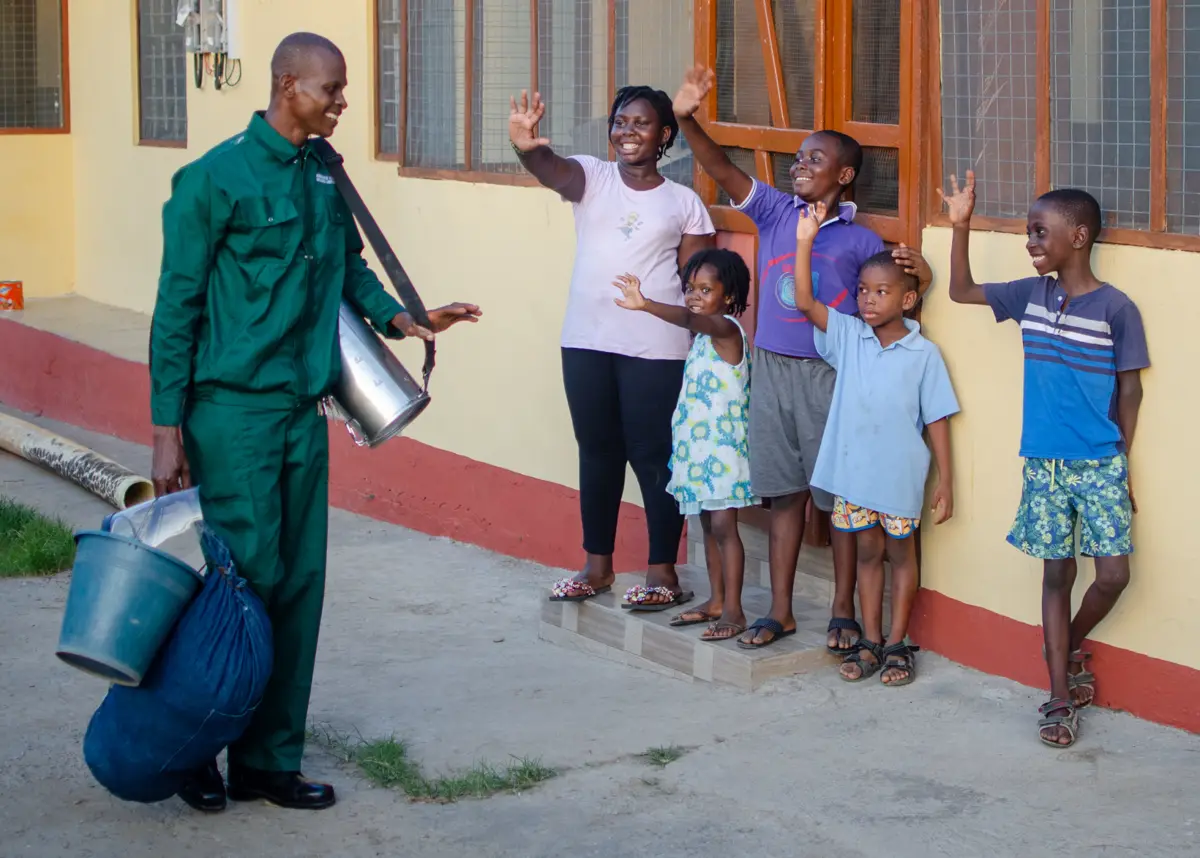A spray operator in green overalls treats the outside wall of a house while children watch